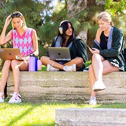 three Chapman female students sitting on a bench looking at their devices